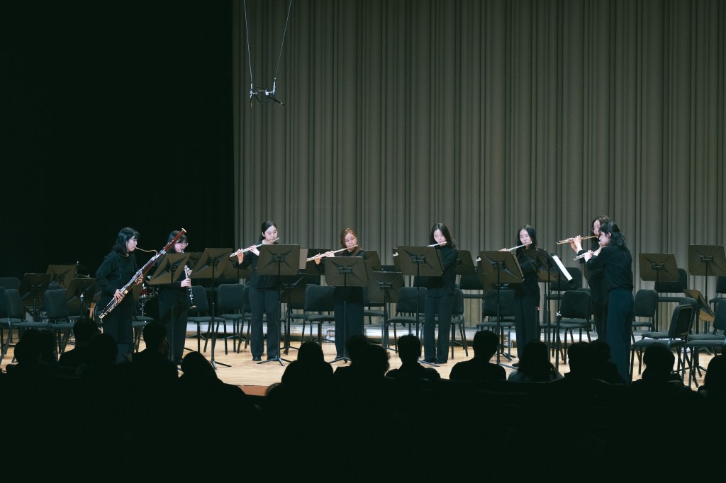 Youth standing in a semi circle on stage playing flutes, clarinets, and bassoon. 